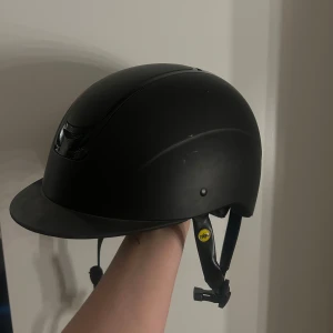 Svart ridhjälm från HH Collection - Säljer en stilren svart ridhjälm från HH Collection. Hjälmen har en slät yta med ventilationsöppningar framtill och justerbart hakband för optimal passform. Perfekt för ridning. Den här varit använd men inget som märks så mycket. 