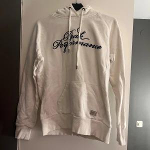 Vit hoodie från Peak Performance - Säljer en vit hoodie från Peak Performance med blå text på bröstet. Den har en klassisk känguruficka och justerbar huva med snören. Perfekt för en avslappnad stil. Har en liten fläck under texten 