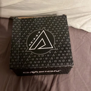 Elektronisk enhet i kartong - En kartong märkt med 'Dvision' och 'Nosai' innehåller en elektronisk enhet med flera anslutningar och fjärrkontroller. Enheten är delvis insvept i bubbelplast för skydd.