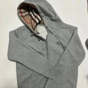 Burberry kofta - Säljer en stilren grå hoodie från Burberry med dragkedja och klassiskt rutigt foder i huvan. Tröjan har en broderad logga på bröstet och ribbade muddar vid ärmslut och nederkant. Perfekt för en avslappnad och trendig look.