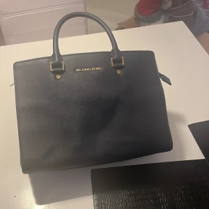 Mörkblå väska från Michael Kors 34cm lång - Snygg svart handväska från Michael Kors i skinn med guldfärgade detaljer. Väskan har två handtag och en dragkedja upptill. Perfekt för att bära dina dagliga nödvändigheter med stil.