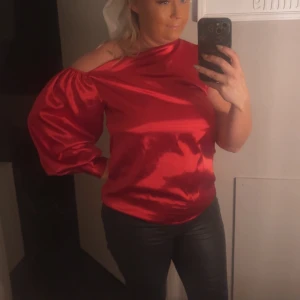 Röd satinblus med oneshoulder-design - Snygg röd satinblus med en unik oneshoulder-design och puffig lång ärm. Perfekt för en kväll ute eller en speciell tillställning. Blusen har en glansig finish som ger en lyxig känsla.