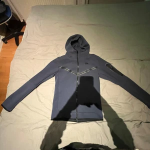 Mörkblå hoodie med dragkedja - Säljer en stilren mörkblå hoodie med dragkedja och huva. Tröjan har långa ärmar och en liten logga på bröstet. Perfekt för en avslappnad look.