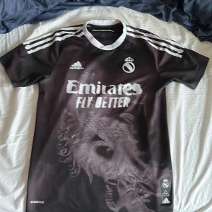 Humanrace real Madrid tröja - Snygg svart fotbollströja från Adidas med vit text och detaljer. Tröjan har korta ärmar och ett coolt lejonmönster på framsidan. 'Humanrace' står tryckt på både fram- och baksidan. Perfekt för fotbollsfans som vill sticka ut!
