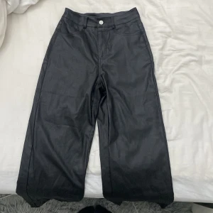 Läderbyxor  - Snygga svarta byxor i ett glansigt material med hög midja och vida ben. Perfekta för en trendig look. Byxorna har fickor både fram och bak samt en knapp och dragkedja framtill.