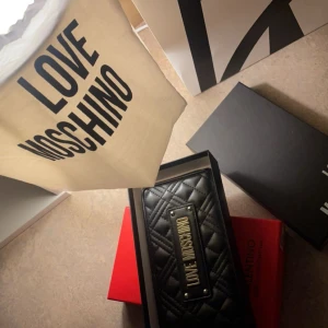 Svart quiltad plånbok från Love Moschino - Snygg svart quiltad plånbok från Love Moschino med dragkedja och en elegant logotypplatta i metall. Perfekt för att hålla dina kort och kontanter organiserade med stil.boxen och dustbagen ingår