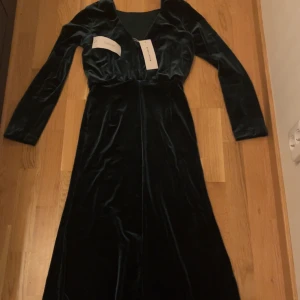 Mörkgrön sammetklänning från Bubbleroom - Elegant mörkgrön långklänning i sammet från Bubbleroom. Klänningen har en v-ringad design och långa ärmar, perfekt för höst- och vinterevenemang. Den mjuka sammeten ger en lyxig känsla.