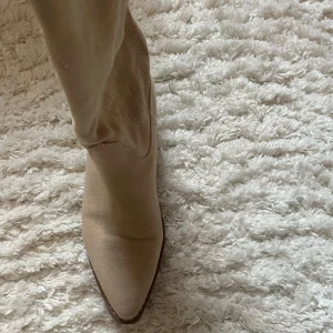 Beige cowboy boots med broderad detalj - Snygga beige cowboy boots i mocka med broderade detaljer. De har en låg klack och spetsig tå, perfekt för en stilren look.