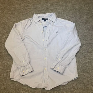 Ljusblå skjorta från Ralph Lauren - Säljer en stilren ljusblå skjorta från Ralph Lauren med klassisk krage och broderad logotyp på bröstet. Skjortan har långa ärmar och knappar framtill. Perfekt för en elegant look. Storlek 18 som motsvarar S/XS