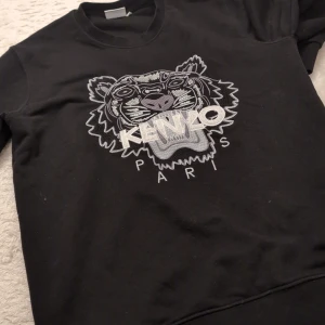 Svart sweatshirt från Kenzo  - Snygg svart sweatshirt från Kenzo med ett broderat tigerhuvud och texten 'Kenzo Paris' på framsidan. Tröjan har långa ärmar och en rund halsringning. Perfekt för en stilren och avslappnad look. DISKUTERA PRIS