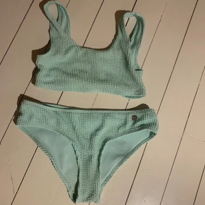 Mintgrön bikini från Kappahl - Snygg mintgrön bikini från Kappahl med en ribbad textur. Toppen har breda axelband och byxorna har en liten metallknappdetalj. Perfekt för sommarens badäventyr! Storlek 146-152 barn men den är stretchig så kan användas av 170. Nypris: 300kr