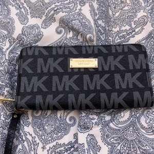 Svart plånbok från Michael Kors - Snygg svart plånbok från Michael Kors med MK-logga och guldfärgad metallplatta. Plånboken har flera fack och en dragkedja för säker förvaring. Perfekt för att hålla ordning på kort och kontanter.