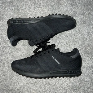 Svarta Adidas LA Trainer sneakers - Snygga svarta Adidas LA Trainer sneakers med klassisk design. Skorna har en stickad ovandel och snörning för en bekväm passform. Perfekta för dig som vill uppnå en casual stil. Tveka inte på att höra av er vid några som helst funderingar