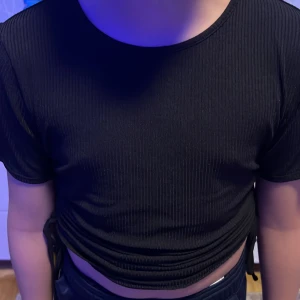 Svart ribbad croppad topp - Snygg svart ribbad topp med korta ärmar och croppad passform. Toppen har knytning i sidan för en justerbar look. Perfekt för en trendig stil.