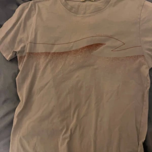 Grå t-shirt från Nudie Jeans - Säljer en vit t-shirt från Nudie Jeans med ett stilrent tryck i rött över bröstet. Perfekt för en avslappnad stil. T-shirten har en klassisk rund halsringning.