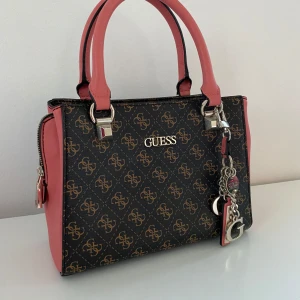 Guess Väska  - Snygg handväska från Guess i brun med rosa detaljer och guldiga logotyper. Väskan har två handtag och en dekorativ kedja med Guess-berlocker. Den har flera fack med dragkedja för praktisk förvaring. Nyttskick.