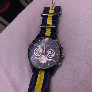 Blå klocka med randigt armband från Gant - Snygg klocka från Gant med blå urtavla och flera sub-dials. Armbandet är randigt i blått och gult, vilket ger en sportig look. Perfekt för dig som vill ha en stilren och funktionell accessoar.