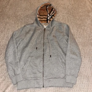 Grå zip hoodie Burberry - Säljer en stilren grå hoodie från Burberry med dragkedja och klassiskt rutigt mönster i huvan. Tröjan har en broderad logotyp på bröstet och ribbade muddar vid ärmslut och nederkant. Perfekt för en avslappnad och trendig look.