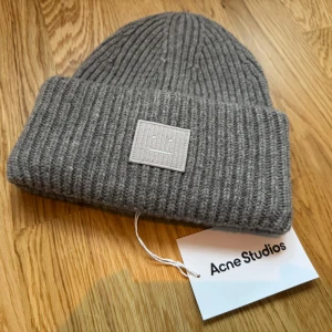 Acne Studios Large Face Logo Beanie NY - Helt ny och oanvänd. Snygg och stilren mössa från Acne Studios. Mössan är i grått och har en klassisk design. Perfekt för kalla dagar.