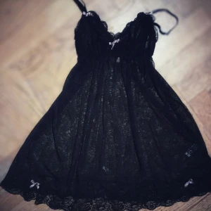 Svart baby doll i spets med rosetter - Elegant svart baby doll klänning i spets med tunna axelband och söta rosetter. Klänningen har en vacker spetsdesign och en feminin siluett. Perfekt för en speciell kväll.