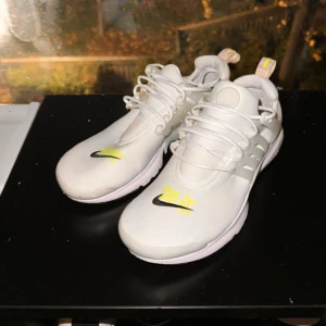 Vita Nike sneakers med gul detalj - Snygga vita Nike sneakers med en gul detalj och svart Nike-logga. Skorna har snörning och en modern design som passar perfekt för en sportig look. Knappt använd