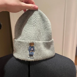 Grå ribbad mössa från Polo Ralph Lauren - Snygg grå ribbad mössa från Polo Ralph Lauren med en broderad nalle på framsidan. Perfekt för kyliga dagar och ger en stilren look.