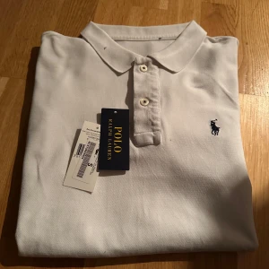 Vit pikétröja från Ralph Lauren - Säljer en klassisk vit pikétröja från Ralph Lauren med den ikoniska broderade loggan på bröstet. Tröjan har en krage och knappar framtill, perfekt för en stilren look. Passar både till vardags och mer uppklädda tillfällen. Ny pris 1599kr ingen bruksmerker