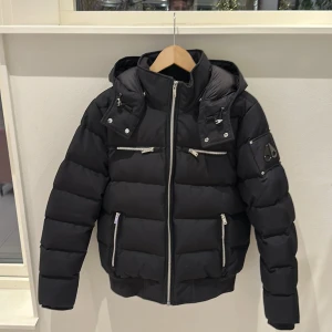 Svart pufferjacka från Moose Knuckles - Snygg svart pufferjacka från Moose Knuckles med huva och flera dragkedjefickor. Jackan har en quiltad design och är perfekt för kalla dagar. Den har en cool logga på ärmen och en bekväm passform. Kvitto finns!!! Bara fråga andvänd måttligt 