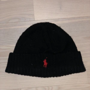 Svart mössa från Ralph Lauren - Snygg svart mössa från Ralph Lauren med röd broderad logga framtill. Perfekt för kyliga dagar med sin ribbade kant som ger en skön passform. Bra skick, fraktar direkt. Inga defekter såklart.