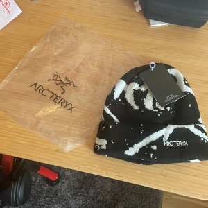 Svart och vit mössa från Arc'teryx - Snygg svart och vit mössa från Arc'teryx med ett abstrakt mönster. Perfekt för kyliga dagar med stil. Märkesloggan är broderad på framsidan.