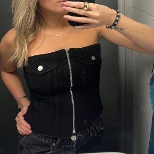 Svart korsett med dragkedja - Snygg svart korsett med dragkedja framtill och dekorativa knappar. Axelbandslös design som ger en stilren look. Perfekt för en kväll ute eller att styla med en kavaj.