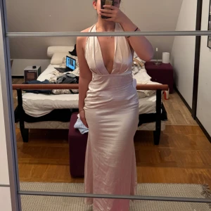 Champagnefärgad långklänning - Elegant champagnefärgad långklänning med djup v-ringning och öppen rygg. Klänningen har en halterneck-design och är tillverkad i ett glansigt material som ger en lyxig känsla. Perfekt för speciella tillfällen.