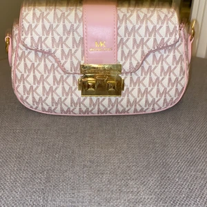 Axelväska från Michael Kors - Snygg axelväska från Michael Kors med monogrammönster i beige och rosa. Väskan har en justerbar axelrem i rosa och en elegant guldfärgad låsdetalj framtill. Perfekt för att ge din outfit en lyxig touch.