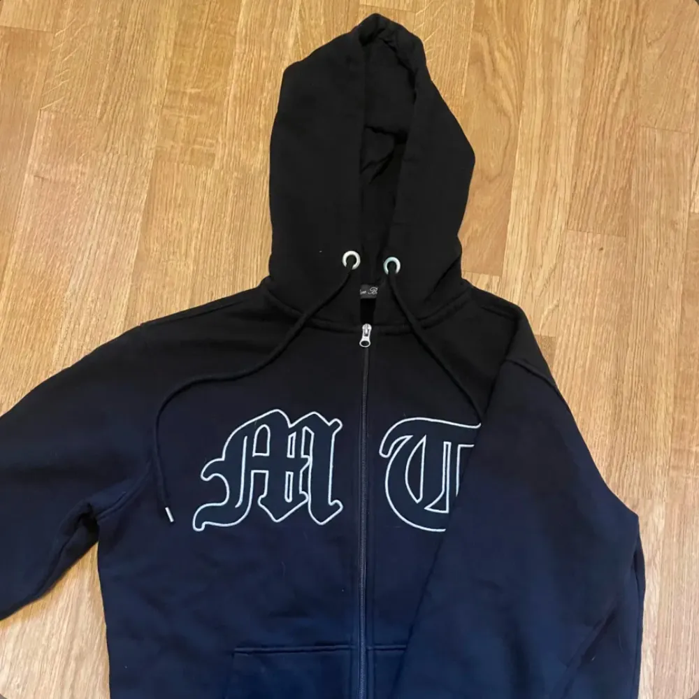 Snygg svart hoodie med dragkedja från Sinners Attire. Den har en stor huva och tryck med bokstäverna 'MT' på framsidan. Perfekt för en casual look.. Neuletakit & Villapaidat.