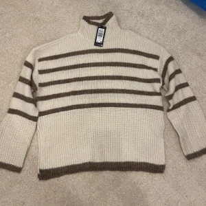 Beige och brun randig stickad tröja från Vero Moda - Helt ny och oanvänd med prislappen kvar. Mysig stickad tröja i beige med bruna ränder. Tröjan har en hög krage och långa ärmar. Perfekt för kyliga dagar. Skriv för mer information 🩷