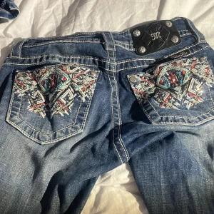 Blå jeans med broderade bakfickor från Miss Me - Snygga blå jeans från Miss Me med unika broderade detaljer på bakfickorna. Jeansen har en klassisk femficksdesign och en dekorativ knappstängning. Perfekta för dig som vill ha något extra i garderoben!