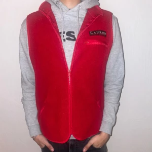 Röd fleeceväst från Lauren - Säljer en snygg röd fleeceväst från Lauren med dragkedja framtill. Västen har en broderad logga på bröstet och är perfekt för lager-på-lager-stil. Passar bra över en hoodie eller tröja.