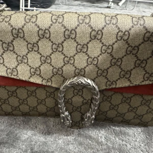 Beige och röd axelväska med monogram - Snygg axelväska med klassiskt monogrammönster i beige och rött. Väskan har en elegant silverkedja och ett dekorativt spänne framtill. Perfekt för att ge din outfit en lyxig touch.