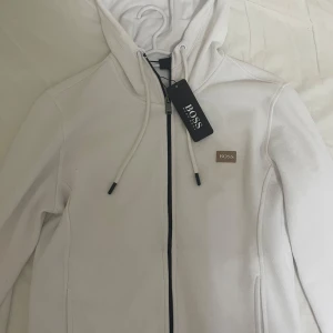 Vit huvtröja från BOSS - Snygg vit huvtröja från BOSS med dragkedja och justerbar huva. Perfekt för en stilren och avslappnad look. Märkeslogga på bröstet ger en exklusiv touch.