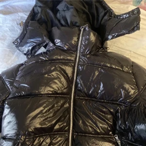 Svart glansig pufferjacka från 157 - Snygg svart pufferjacka från 157 med glansig yta och dragkedja framtill. Jackan har en hög krage och är perfekt för kyligare väder. Den är designad för att hålla dig varm och stilren. BYTER GÄRNA MOT EN SMALL!!!! 
