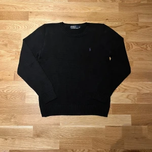 Svart stickad tröja från Polo Ralph Lauren - Säljer en snygg svart stickad tröja från Ralph Lauren med deras ikoniska logga broderad i lila på bröstet. Tröjan har en rund halsringning och långa ärmar med ribbade muddar. Passar lite mer som S. Perfekt för en klassisk look.