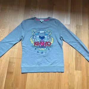 Grå sweatshirt från Kenzo - Säljer en grå sweatshirt från Kenzo med ett färgglatt tigerbroderi på framsidan. Tröjan har långa ärmar och en rund halsringning med rosa detaljer på insidan. Perfekt för en avslappnad stil.