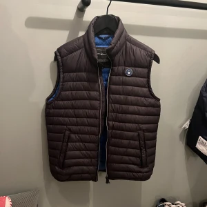 Svart dunväst från Sail Racing - Snygg svart dunväst med blå insida och dragkedja. Västen har en hög krage och ett runt märke på bröstet. Perfekt för lager-på-lager under kyliga dagar.