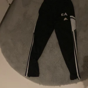 Svarta träningsbyxor från Adidas - Säljer ett par svarta träningsbyxor från Adidas med vita ränder längs sidorna och EA-logga. Perfekta för sport och träning. Byxorna har en bekväm passform och elastisk midja.