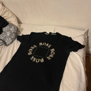 Svart t-shirt från Boss - Snygg svart t-shirt från Boss med ett cirkulärt tryck av logotypen på framsidan. Perfekt för en stilren look. Kortärmad och tillverkad i mjukt material för bekväm passform. Äkta QR Scan finns. Skriv för intresse 