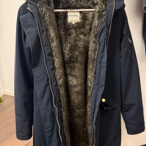 Mörkblå parkajacka från Elvine - Snygg mörkblå parkajacka från Elvine med fuskpälsfoder. Jackan har dragkedja och knappar framtill samt praktiska fickor. Perfekt för kyliga dagar.