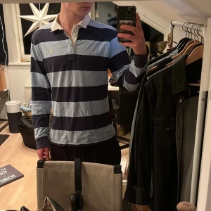 Randig pikétröja från Ralph Lauren - Säljer en klassisk randig pikétröja från Ralph Lauren i blå och ljusblå. Tröjan har långa ärmar och en kontrasterande krage i beige och rosa. Perfekt för en stilren look. Storlek m 