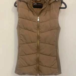 Brun dunväst från Zara - Snygg beige dunväst från Zara med dragkedja och huva. Perfekt för kyliga dagar när du vill hålla dig varm och stilren. Säljer min bruna väst från zara på grund av få användning. Ett hänge på ena fickan har lossnat men det syns nästan inte eftersom den är så pass liten och fickan funkar ändå. Köparen står för frakten