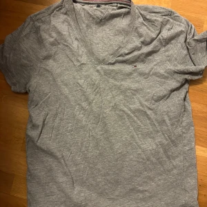 Grå t-shirt från Hilfiger Denim - Säljer en grå t-shirt från Hilfiger Denim med v-ringning. Den har en liten broderad logga på bröstet och är kortärmad. Perfekt för en avslappnad stil.
