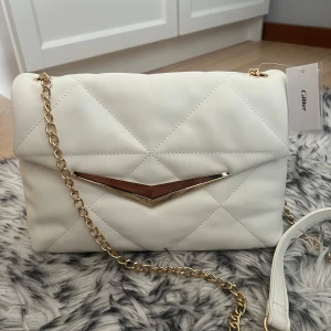 Vit axelväska med gulddetaljer - Säljer en superfin vit oanvänd axelväska med gulddetaljer med tillhörande guldkedja. Passar perfekt både till vardags och fest för att piffa upp outfiten. Ny pris:299kr